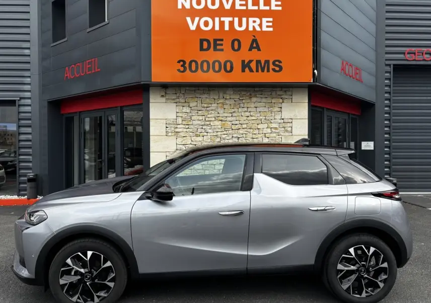 Vue latérale droite d'une DS3 2024 gris clair avec toit noir, jantes noires et détails modernes devant un bâtiment commercial.