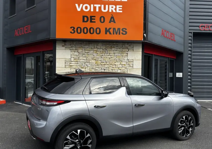 Vue latérale droite d'une DS3 2024 gris clair avec toit noir, jantes alliage noires et vitres teintées, garée devant un bâtiment.