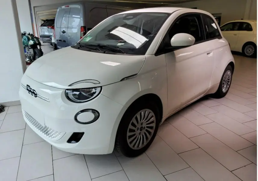 Vue 3/4 avant droite d'une Fiat 500 électrique blanche stationnée en intérieur avec jantes spécifiques et phares ronds LED.