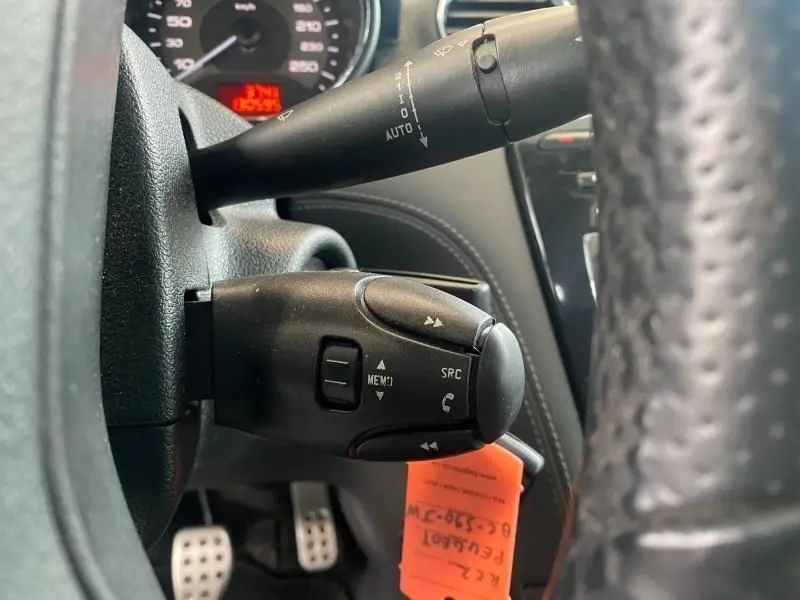 Gros plan sur les commandes audio au volant noir du Peugeot RCZ gris, avec tableau de bord et pédales visibles en arrière-plan.