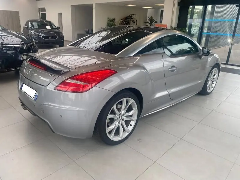 Peugeot RCZ gris vue 3/4 arrière droit en showroom, avec jantes alliage et aileron arrière déployé.