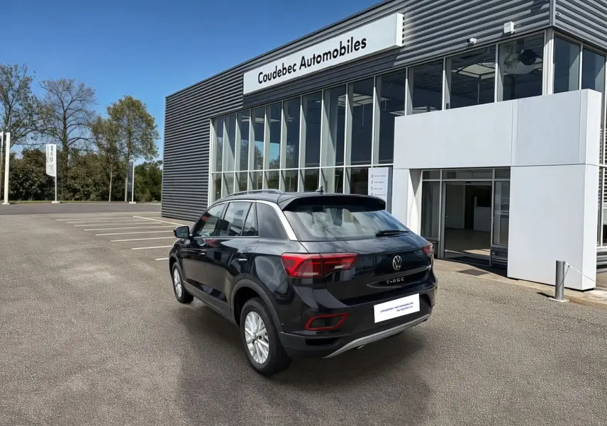 Vue 3/4 arrière droite d'un Volkswagen T-Roc noir 2025 stationné devant un bâtiment moderne de concession.