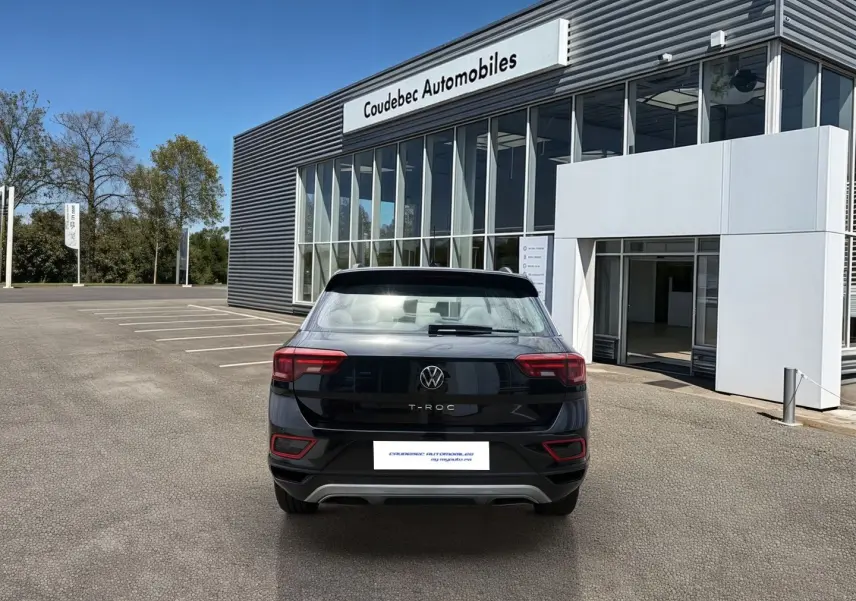 Vue arrière d’un Volkswagen T-Roc noir 2025 stationné devant un bâtiment moderne de concession automobile.