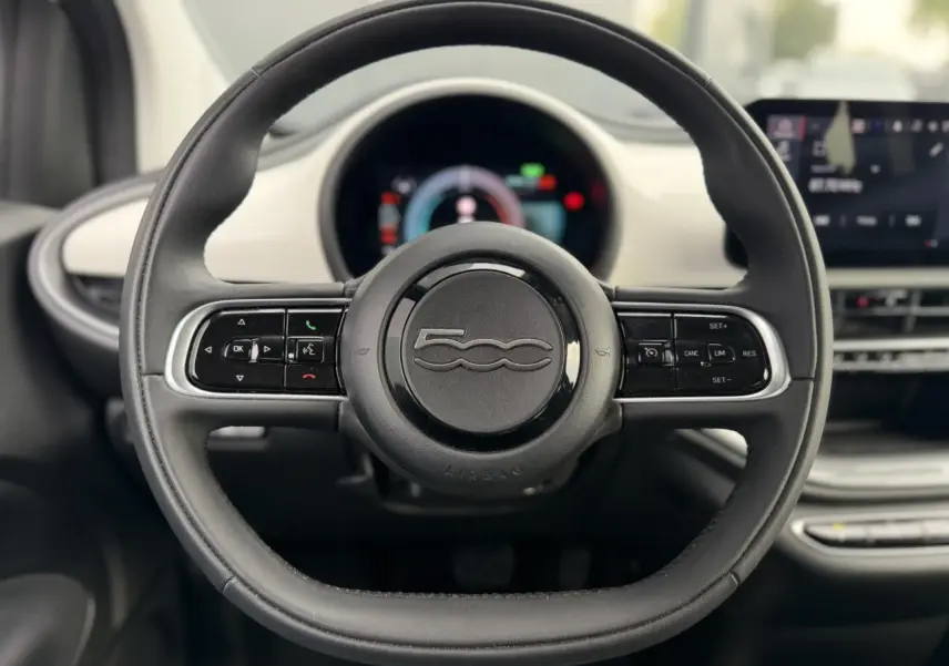 Vue centrée sur le volant noir de la Fiat 500 électrique 2024 avec tableau de bord numérique en arrière-plan.
