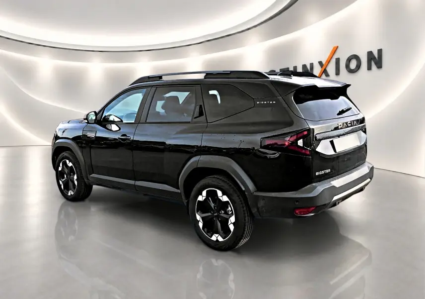 Vue 3/4 arrière droite du SUV Dacia Bigster noir avec jantes alu et barres de toit dans un showroom lumineux.
