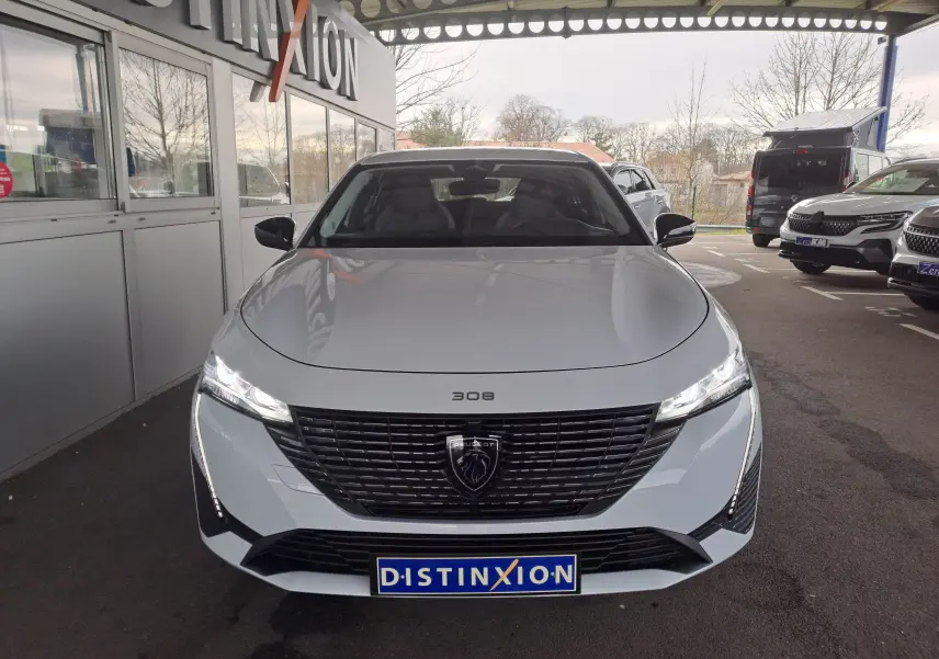 Vue frontale d'une Peugeot 308 blanche Okenite avec calandre noire et phares LED allumés sous un abri.