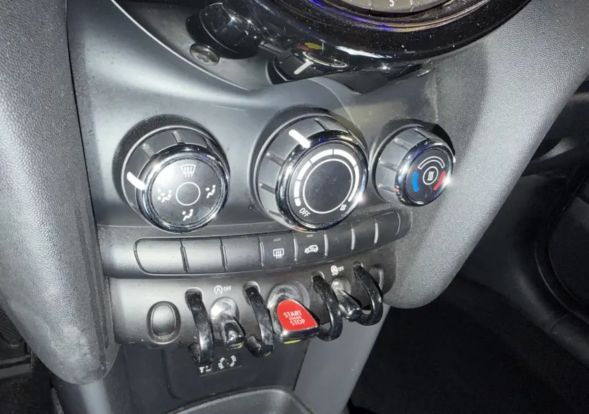 Vue rapprochée du tableau de bord noir de la MINI 1.5i 75CV ONE, avec commandes de climatisation et bouton start/stop rouge.