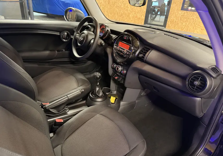 Vue intérieure côté conducteur de la MINI 1.5i noire, avec tableau de bord rond et levier de vitesses manuel visible.