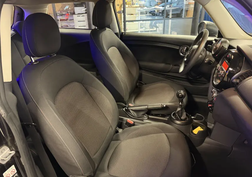 Vue intérieure côté gauche de la MINI 1.5i noire, montrant les sièges avant, volant, et levier de vitesse manuel.
