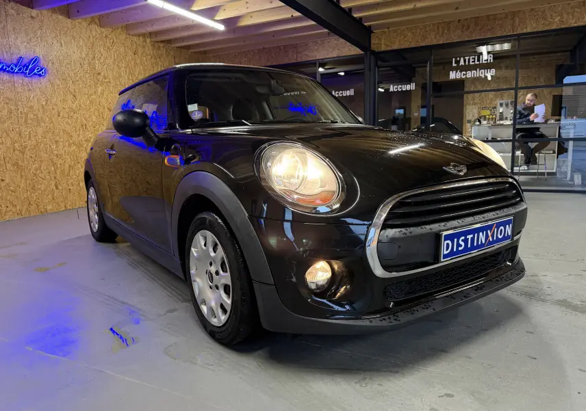 MINI noire vue en 3/4 avant droit, phares et antibrouillards allumés, dans un atelier au sol gris.