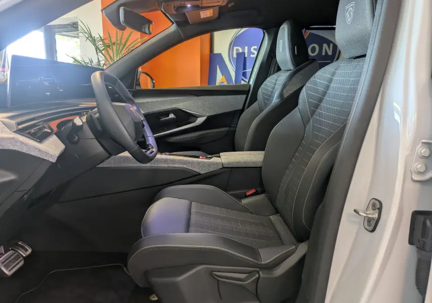 Vue intérieure côté conducteur du Peugeot 5008 Hybrid 145 e-DCS6 GT 2025, sièges tissu/tepp noir avec surpiqûres claires et volant multifonction.
