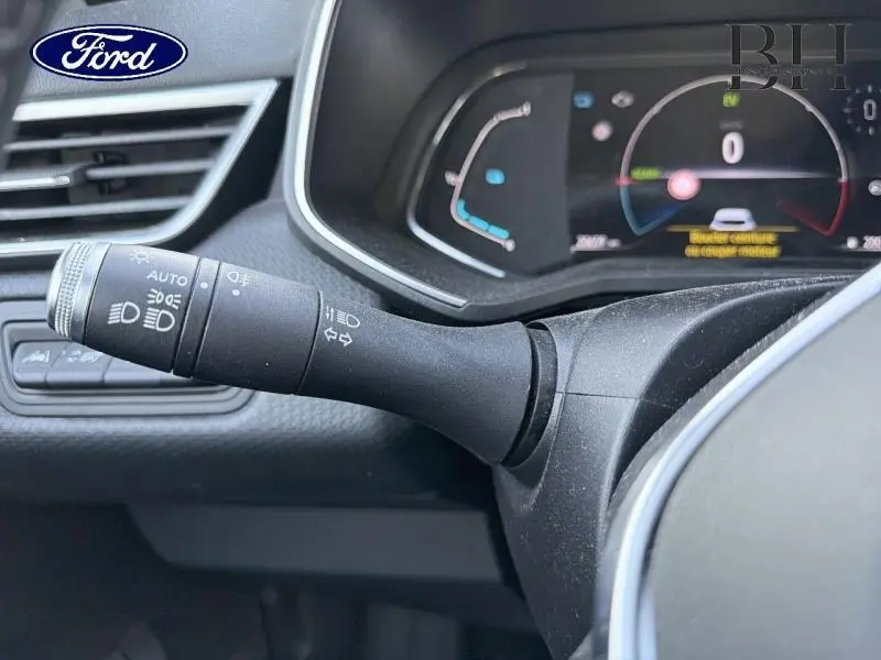 Le véhicule visible est une Renault Clio 1.6 E-Tech blanche, mais l'image montre uniquement un détail intérieur du levier de commande des phares et clignotants côté gauche du volant, avec le tableau de bord numérique en arrière