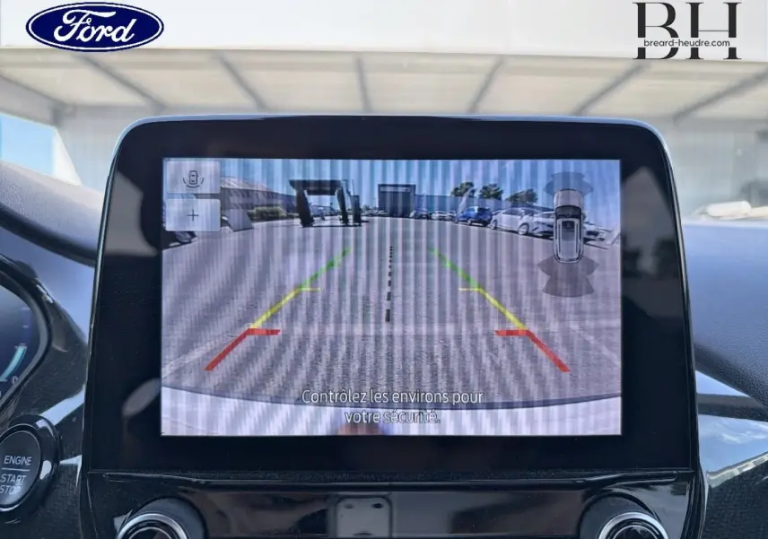 Écran tactile intérieur montrant la caméra de recul avec vue arrière sur un parking et véhicules stationnés.