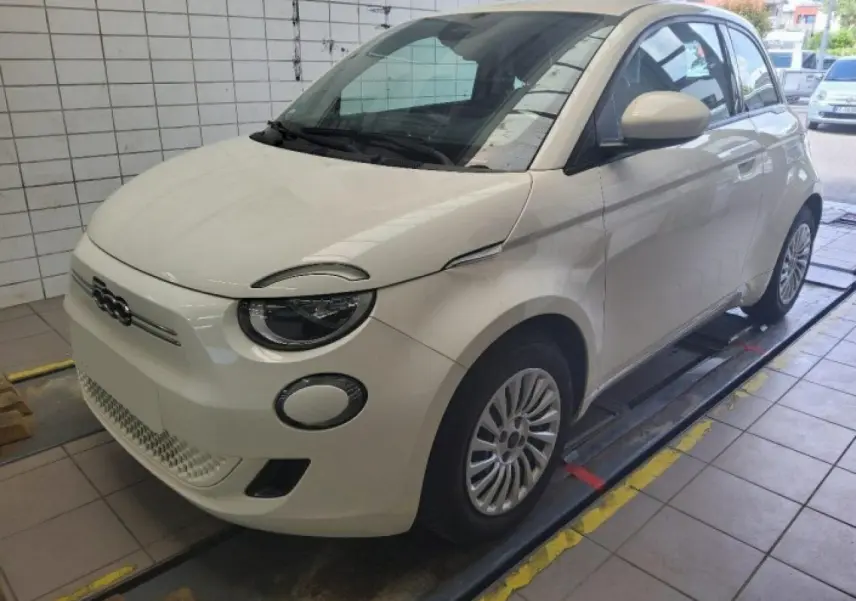 Vue 3/4 avant droite d'une Fiat 500 électrique blanche stationnée dans un garage avec phares ronds distinctifs.