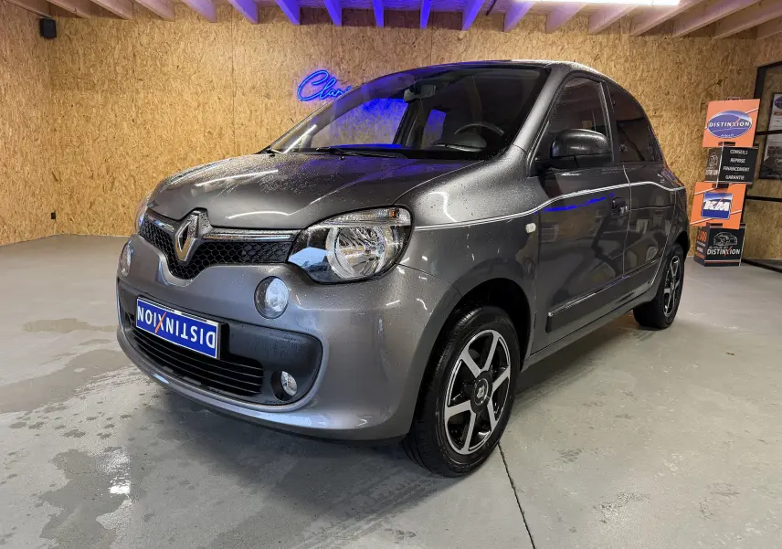 Renault Twingo III gris en 3/4 avant droit, avec jantes noires et phares halogènes visibles en intérieur showroom.