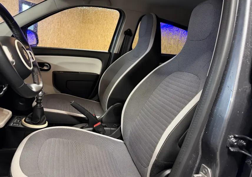 Vue intérieure côté conducteur de la Renault Twingo III gris, montrant les sièges avant et le volant cuir avec levier de vitesse manuel.