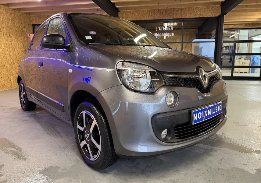 Renault Twingo III gris vue 3/4 avant droit, avec phares halogènes et jantes alliage noires, stationnée en intérieur.