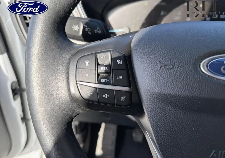Gros plan sur le volant cuir multifonction noir du Ford Kuga blanc Glacier, avec commandes de régulateur et limiteur de vitesse.