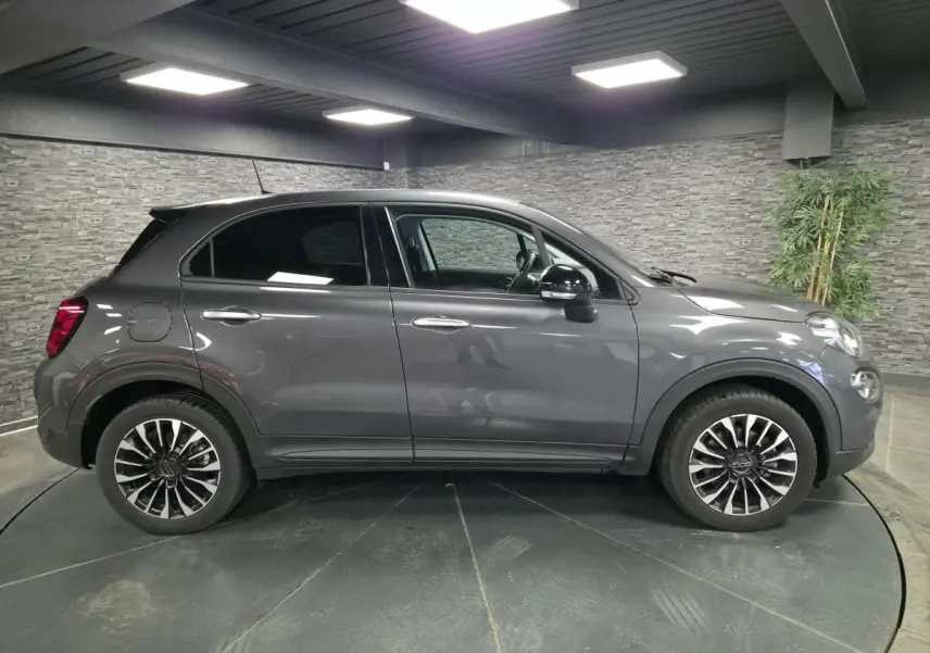 Profil droit d'un Fiat 500X gris métallisé 2024 avec jantes alliage 17 pouces et vitres surteintées dans un showroom.