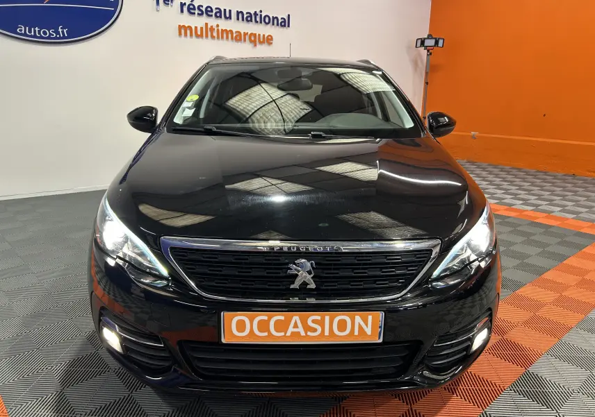 Vue frontale d'une Peugeot 308 SW noire avec phares allumés et plaque "OCCASION" en intérieur showroom.