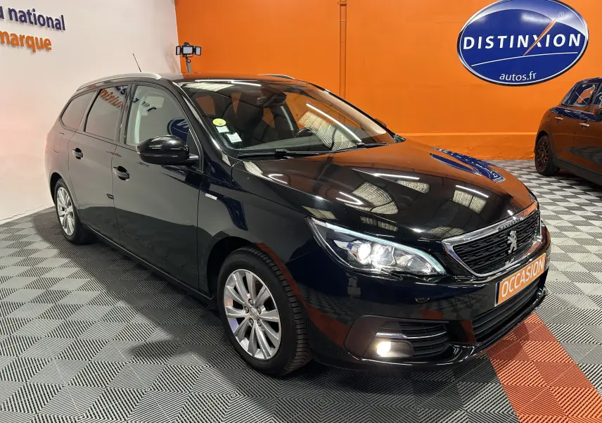 Peugeot 308 SW noire vue en 3/4 avant droit, avec phares allumés et attelage visible au garage.