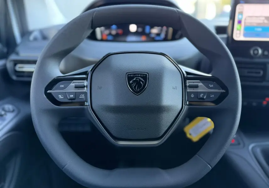 Vue rapprochée du volant multifonctions noir avec logo Peugeot, tableau de bord numérique et écran tactile en arrière-plan.