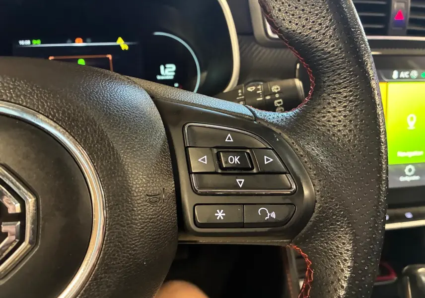 Gros plan sur les commandes à droite du volant en cuir noir perforé avec surpiqûres rouges du MG ZS blanc, tableau de bord en arrière-plan.