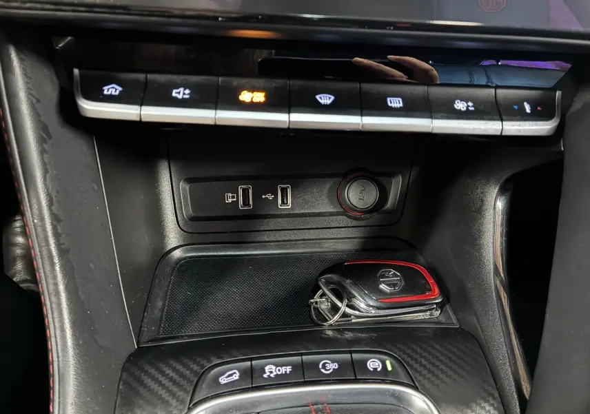 Gros plan sur la console centrale noire avec touches de climatisation, ports USB et clé MG ZS blanche posée.