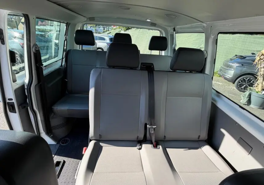 Vue intérieure du Volkswagen Transporter T6 Minibus blanc, montrant les sièges arrière en tissu gris et la porte coulissante ouverte côté gauche.
