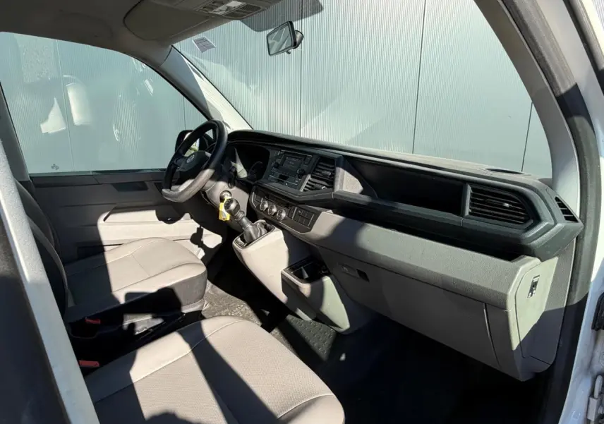 Vue intérieure côté conducteur du Volkswagen Transporter T6 blanc, montrant le tableau de bord et les sièges avant gris.