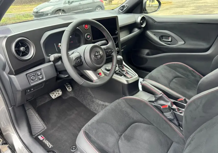 Vue intérieure côté conducteur de la Toyota Yaris GR avec sièges noirs surpiqués rouges et volant sport à coutures rouges.