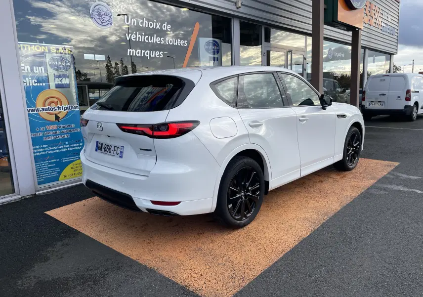Vue 3/4 arrière droite d'un Mazda CX-60 blanc nacré avec jantes noires, garé devant une vitrine de concession.