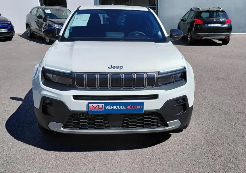 Vue avant d'un Jeep Avenger blanc 2025 avec calandre noire et plaque "Véhicule récent" sur un parking extérieur.