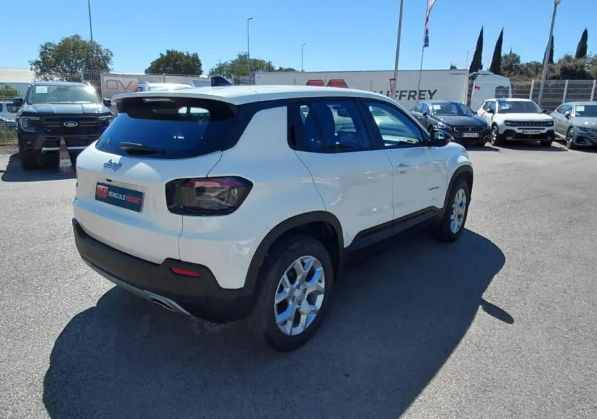Jeep Avenger blanc vu en 3/4 arrière droit sur un parking, avec des jantes alliage et des feux arrière fumés.