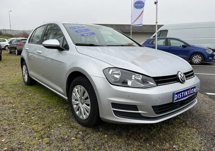 Volkswagen Golf 7 argent vue 3/4 avant droite sous la pluie avec plaques et drapeaux Distinxion visibles.