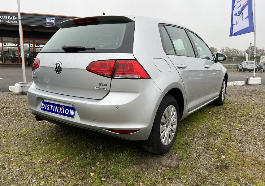 Vue 3/4 arrière droite d'une Volkswagen Golf 7 argent avec badge TDI et plaque Distinxion sur un terrain herbeux.