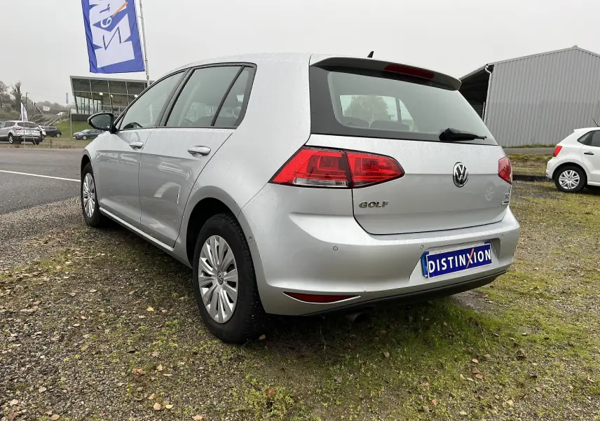 Vue 3/4 arrière droite d'une Volkswagen Golf 7 argentée, garée sur un sol herbeux et gravier, avec feux arrière rouges visibles.