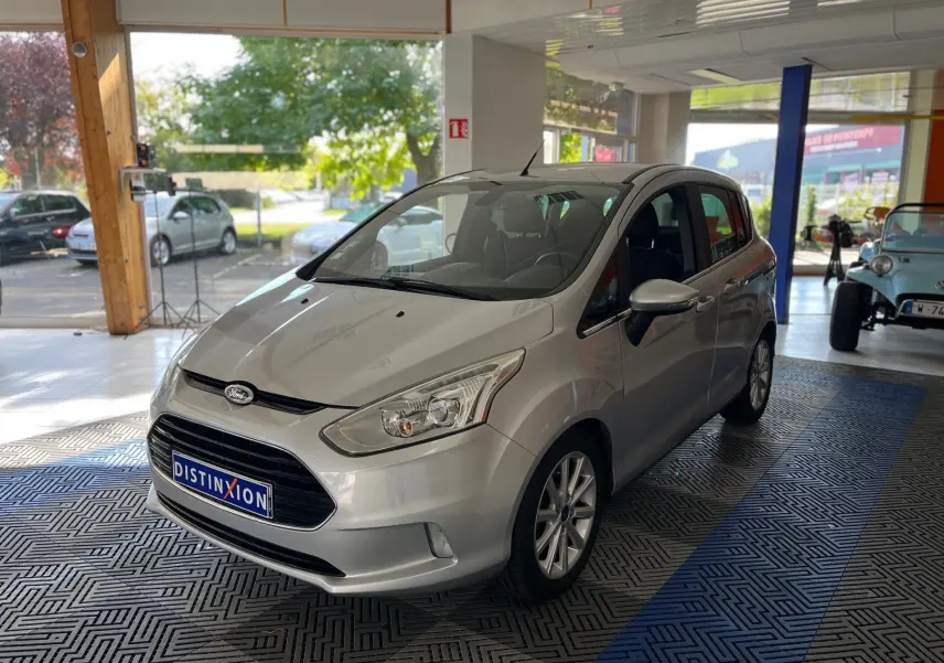 Ford B-MAX gris clair vue 3/4 avant droit en intérieur avec plaque Distinxion visible sur le pare-chocs avant.