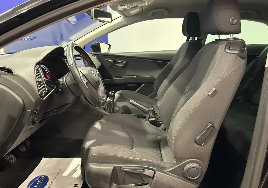 Vue intérieure côté conducteur de la SEAT Leon noire, montrant les sièges avant en tissu et le volant cuir avec levier de vitesses manuel.