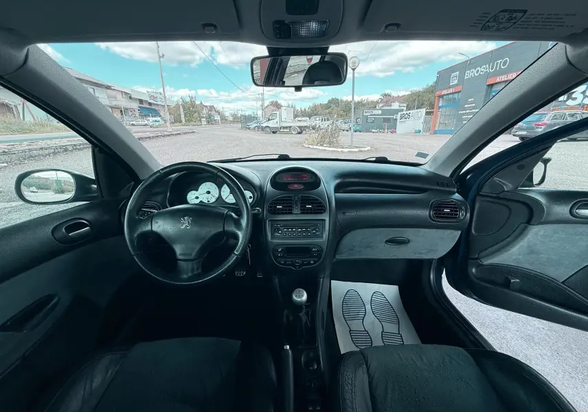 Vue intérieure avant de la Peugeot 206 bleue de 2003, montrant le tableau de bord, volant cuir et levier de vitesse manuel.