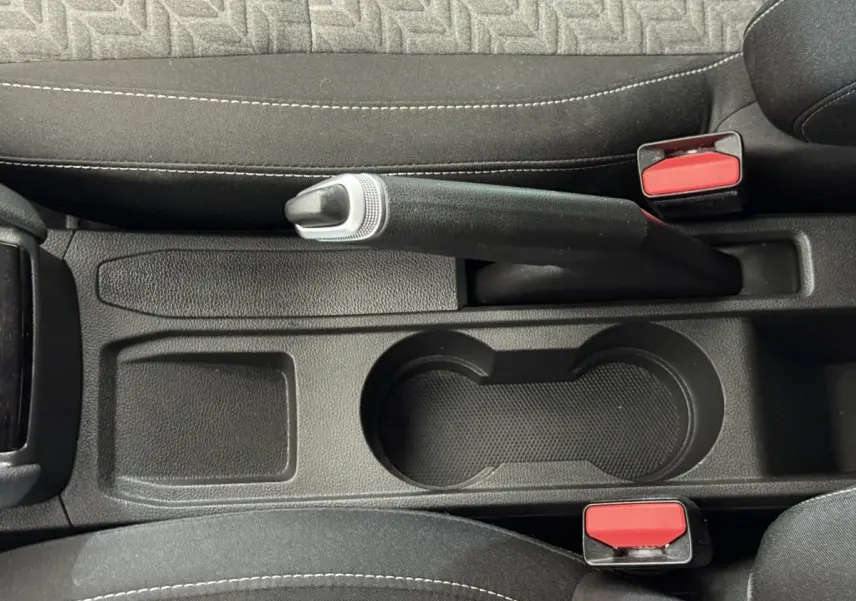 Vue plongeante sur le frein à main et porte-gobelets noirs entre les sièges avant en tissu gris de la Renault Clio blanche.