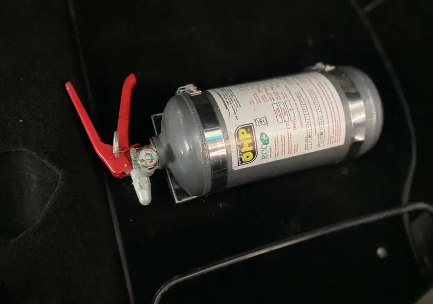 Extincteur OMP gris avec poignée rouge posé sur le sol noir intérieur d'une Porsche 911 3.0 SC 1980.
