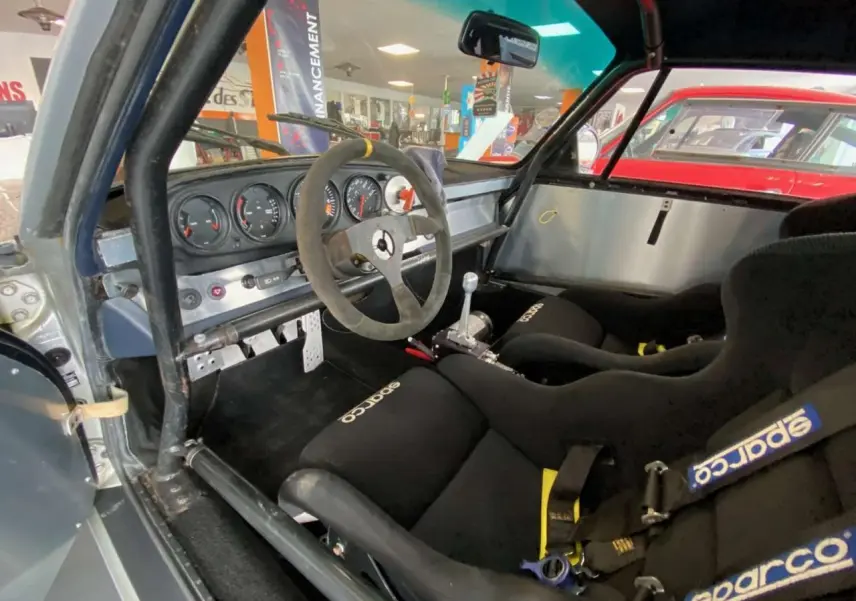 Vue intérieure côté conducteur de la Porsche 911 3.0 SC gris métallisé, avec baquets Sparco et arceau de sécurité.