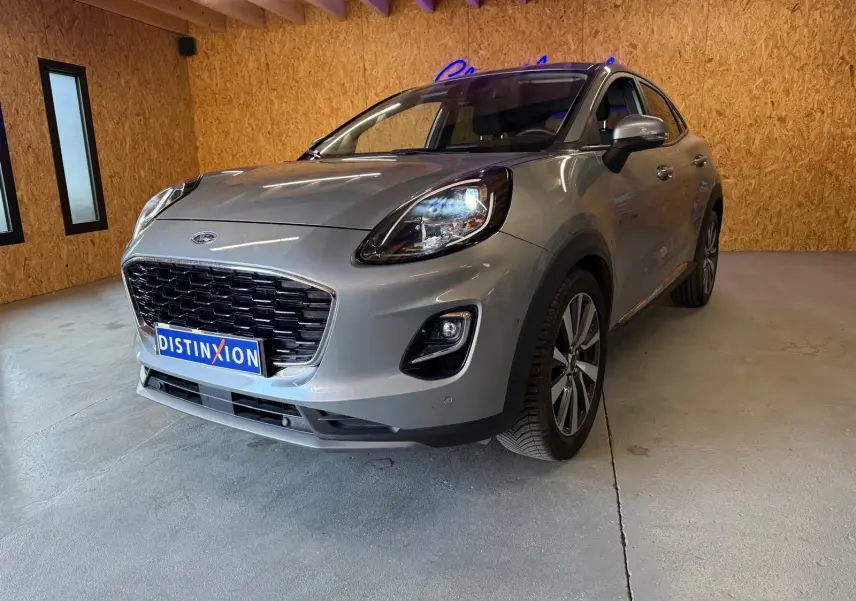 Ford Puma gris en 3/4 avant droit, avec calandre noire et phares allumés dans un garage en bois.