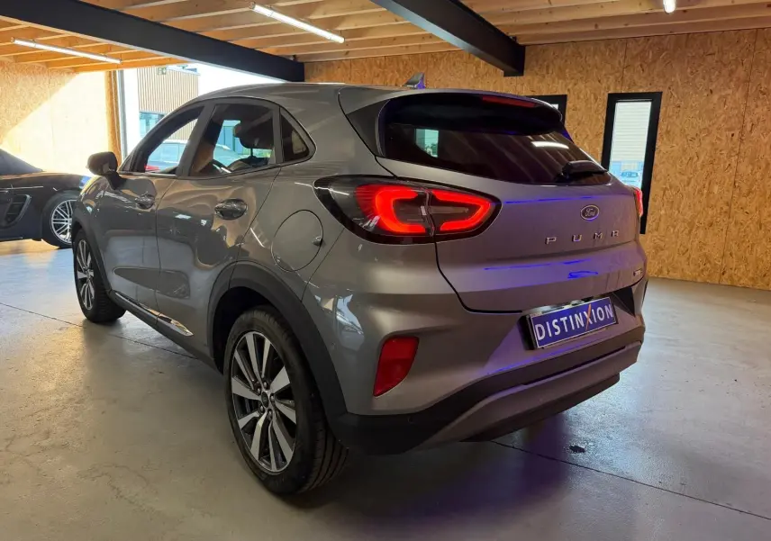 Vue 3/4 arrière droite d'une Ford Puma gris métallisé 2021 avec feux arrière allumés dans un garage lumineux.