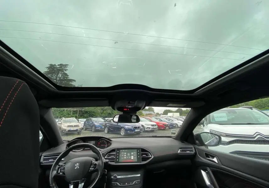 Vue intérieure avant du Peugeot 308 SW bleu magnétique, avec toit panoramique en verre et volant cuir à surpiqûres rouges visible.