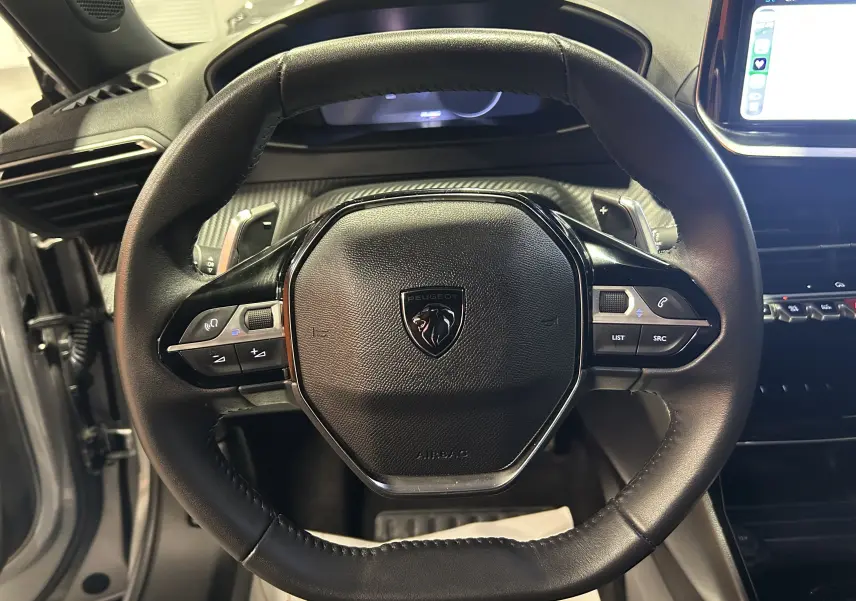 Gros plan sur le volant cuir noir du Peugeot 2008 II Ph.2 Allure Hybrid 145 avec commandes intégrées et tableau de bord numérique.