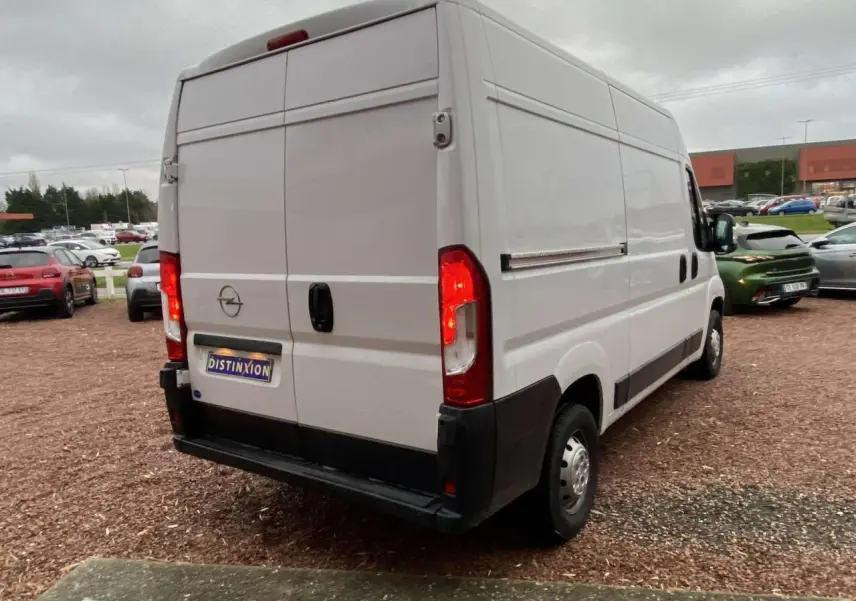 Vue 3/4 arrière droite d’un Opel Movano Fourgon blanc 2022 avec feux arrière allumés sur parking.