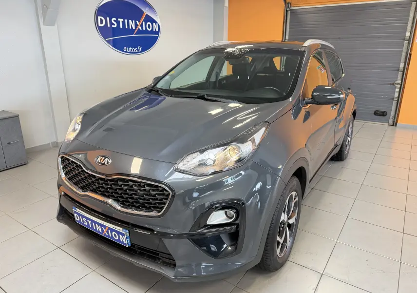 KIA Sportage gris 2019 vue 3/4 avant droit en intérieur, avec calandre noire et phares allumés.