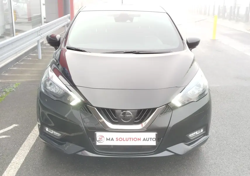 Vue frontale d'une Nissan Micra noire avec phares allumés et calandre chromée, stationnée sur un parking extérieur.